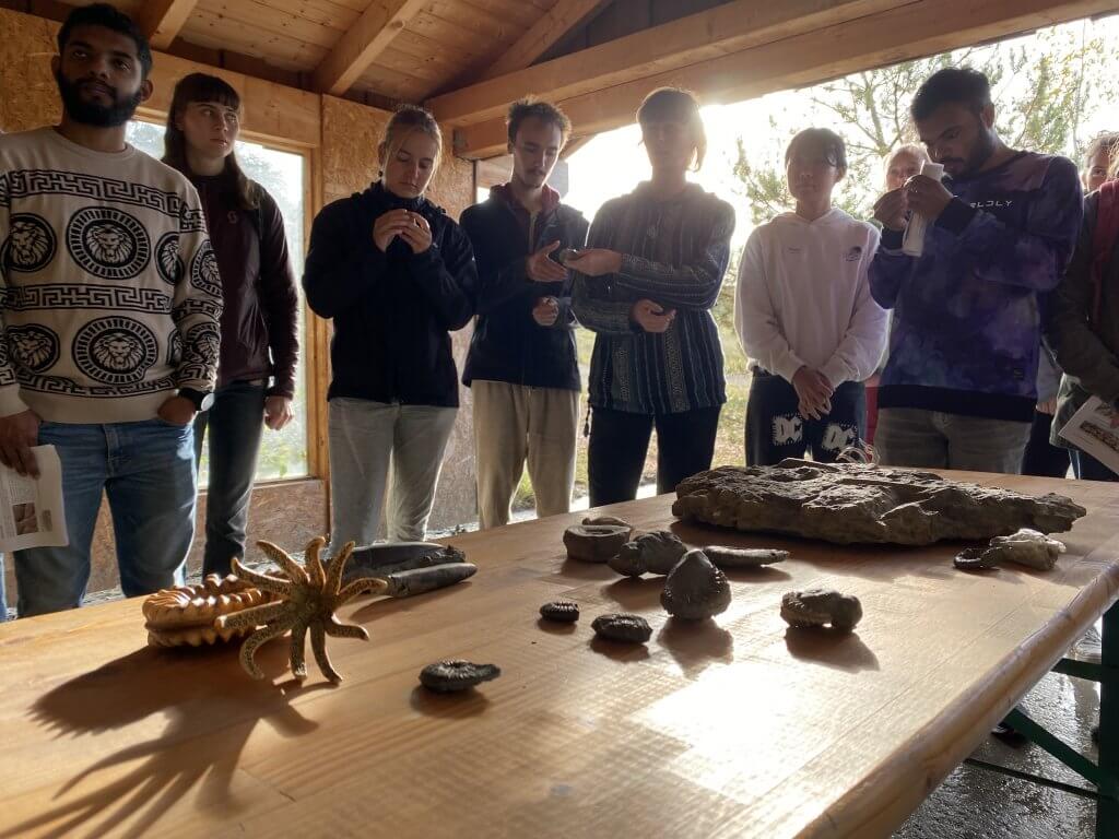 About 10 GCE students standing around a table. On the table are an ammonite model, as well as several fossilised Ammonites and Belemnites. Belemnites have a bullet shape.