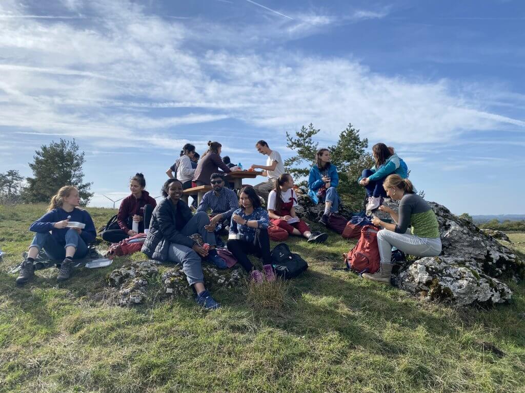A group of students are sitting on rocks in a meadow eating lunch. The sun is shining in the blue sky.